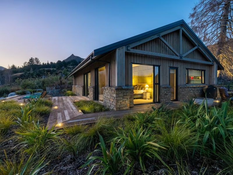 Modern lakeside cabin exterior at dusk, with warm interior lighting and a stone-based deck
