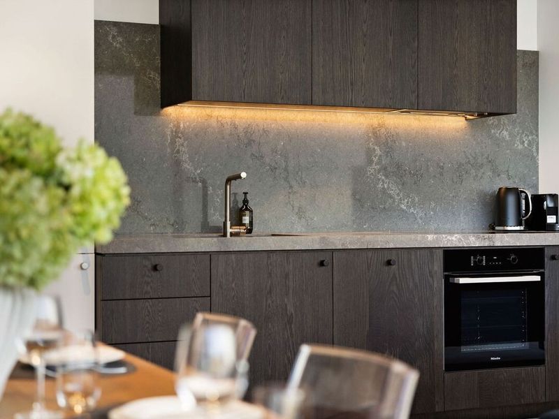 Kitchen counter with dark cabinetry, stone splashback and under-cabinet lighting